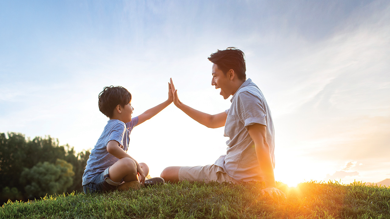 A father and son giving each other a high five on meadow; image used for HSBC Singapore Emerald Life Legacy Plan.