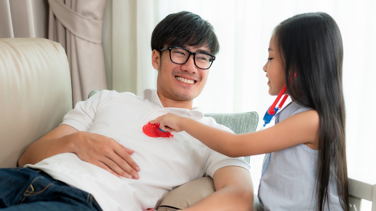 一个女孩用玩具听诊器给父亲听诊；图片使用于汇丰新加坡外籍雇员文章新加坡和其他国家的医疗保险。