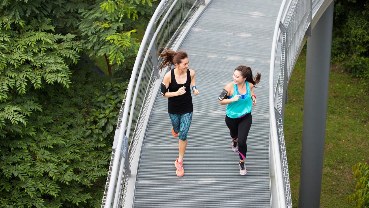两位亚洲女士在绿化带上方的栈道上跑步；图片使用于汇丰“身心健康，乐在当下”页面。